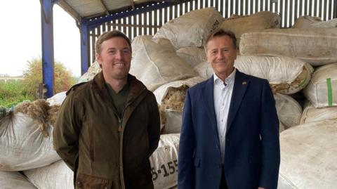 Two men - one wearing a wax coat and the other wearing a suit with open collar - are standing in a barn with bags of wool stacked up behind them. They are both looking towards the camera and smiling.