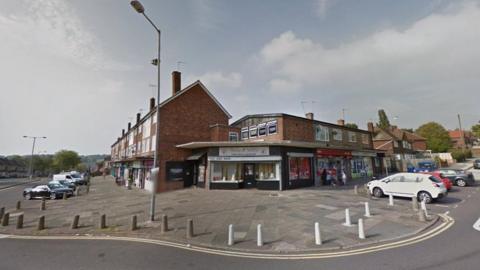 The end of a street with a shop on the corner and other shops on the right hand side. Residential houses surround the shops on either side in a row. The nearby road has a 20mph sign on it. There are cars parked in front of the homes and shops.