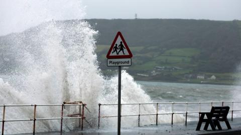 Image of waves crashing against a coastal path. The backdrop behind the waves is a green hill with trees and houses.