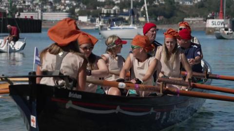 Women wearing cream coloured aprons and orange mob caps are rowing the gig with several men from the lifeboat in the crew too. They are in a harbour.