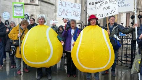 Two women dressed as tennis balls protest outside Royal Courts of Justice with other members of Save Wimbledon Park. They hold various signs with writing such as "Land In Trust For All Of Us!".