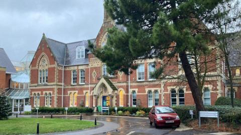 A large red brick Victorian-style building with arched windows and a central entrance, photographed on a wet, overcast day. A red car is parked on the curved driveway in front of the building, with trees and shrubs surrounding the area.