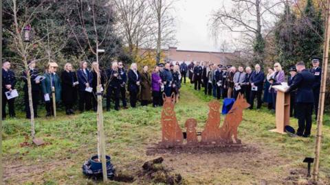 A statue of four different police dogs has been placed in a memorial garden, sapplings have been planted around it. Police officers and staff can be seen paying their respects as an officer gives a reading from a lectern. There are trees and a police building in the background. 