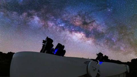 A night sky over several telescopes, black cylinders pointing towards the sky. The sky is brightly lit with stars and ranges in colours from deep blue to white.