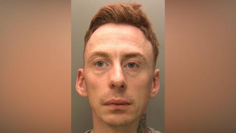 Mugshot of Brian Smith. He has ginger hair which is gelled up and is looking sad and at the camera. He has a neck tattoo of a red rose. He is clean shaven and is wearing a grey jumper. 