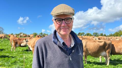 Le Gallais is stood in a field with brown Jersey cows behind him on a sunny day. He is wearing a blue jumper and brown flatcap. He has black glasses.