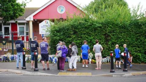 A group of adults and children, seen from behind, look at floral tributes at the entrance to Minehead Middle School in Somerset. Dozens of bunches of flowers have been laid in a row at the bottom of a metal fence.