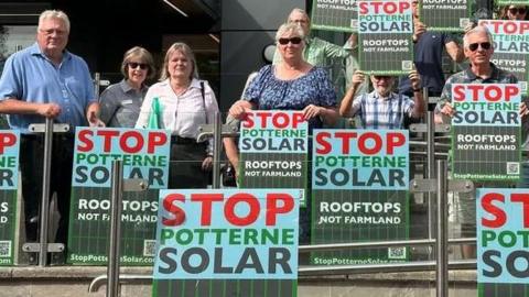 A group of residents holding up "stop potterne solar - rooftops not farmland" signs. They are standing against a glass barrier with a grey office building in the background. 