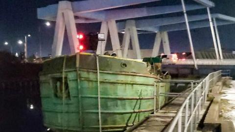 Close-up shot of a barge wedged under a bridge. The barge has a green hull and mooring ropes dangling down. There are railings to the right side of the image.
