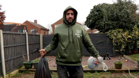 Kyle Newby, wearing a green hoodie, holding a bin bag full of dog poo, stood in a back garden of a house.