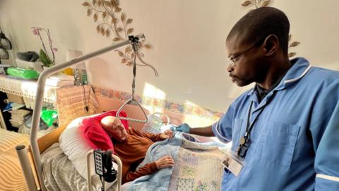 Derek lying in his bed in his living room being cared for by a male carer from Primary Homecare in Suffolk. The carer, called Sandy, is handing him his glasses. Sandy is wearing a blue uniform and a lanyard. Derek is under covers with a grip handle hanging over his bed.