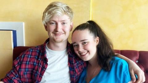 A young couple. He has blond hair and is wearing a tartan shirt with a white T-shirt underneath. She is in a light blue top and has long, dark hair. They are sitting on a red sofa.