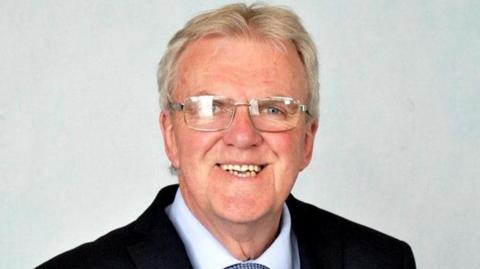 A smiling man with grey hair and silver rimmed glasses in a dark suit with a white tie and a light grey background