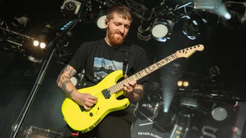 A man with short hair and a beard wears a look of concentration as he plays notes on an electric guitar with a neon yellow body. He's got heavily tattooed arms and the fretboard of his guitar has "It'll B R8 (pronounced rate) printed along its length. 