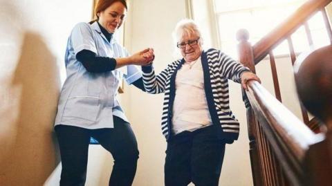 Old woman with grey hair is helped down a stair case by a nurse with a window behind them as sunshine pours through