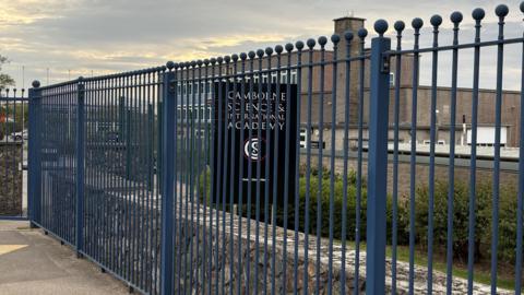 Camborne Science and International Academty school building is in the distance with a black sign with the school's name and logo on it - all behind a blue metal fence. 