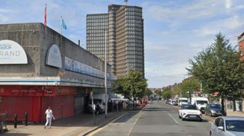 Shot of The Strand shopping centre in Bootle. It is a typical concrete 1970s shopping mall with red shutters in front of the shop on the corner. It is on a tree-lined street with parked cars on either side. In the distance are two high rise buildings. Image from July 2024.