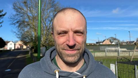 Ben Bonos, 44, has very short cropped hair and stubble on his face, he is wearing a grey hoodie top with a green t-shirt underneath, behind him in a village park with 5-a-side goalposts visible on the right and the new 20m (65ft) tall, green mobile phone signal mast to the left.