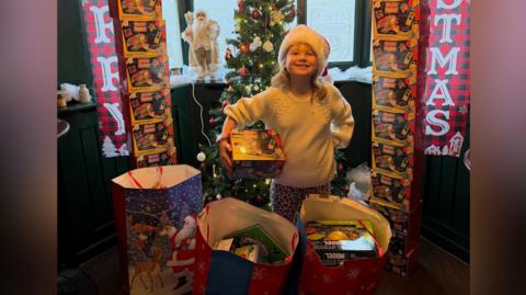 A young girl surrounded by toys. She has one under her arm and she is smiling at the camera. She is wearing a white jumper, leopard legging and a Christmas hat. There is a Christmas tree and other decorations behind her.