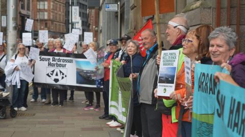People holding banners and placards line the pavement of a Glasgow street. The visible largest banner reads "Save Ardrossan Harbour".