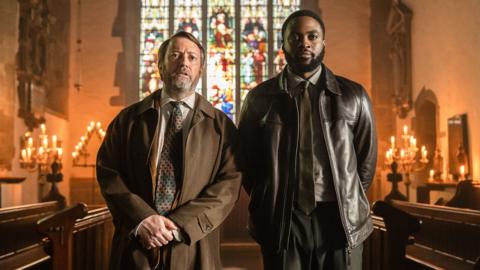 Two men looking at something behind the camera standing in a church with lit candles