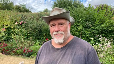 Tim Hoverd wearing a grey hat and dark grey t-shirt is stood in a garden. He has a large hedge with a flower bed behind him. He also has a white beard and is smiling at the camera.