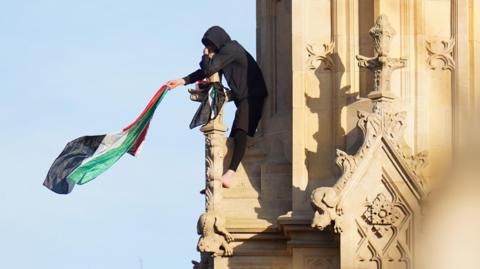 A man holds a Palestinian flag which he waves in the air, as he balances on stonework.