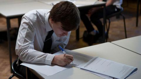 A boy in a white shirt and school tie is sat at a school desk writing on a sheet of paper with a blue biro, with a booklet open in front of him.