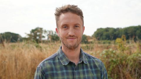 Dominic Buscall, the founder of Wild Ken Hill. He is standing on the nature reserve site in Snettisham, Norfolk, and is looking directly at the camera. He has short blond hair and is wearing a blue and green checked shirt.