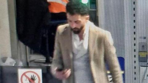 A low-resolution CCTV still image of Ceyhan Dinler walking while looking at his phone. He is wearing a cream jacket and has a white shirt on.
