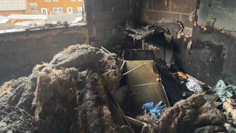 Heavily burned debris, furniture and insulation piled up on the floor of a bedroom. The window has shattered and all the paint and plaster have been stripped from the walls, leaving only blackened breeze blocks.