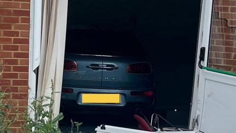 The rear view of a grey car which has crashed through French windows. It has crossed the threshold and is fully into the room. One door is open on the right and the other is in pieces on the ground in front of the brick built building.