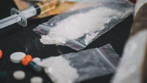 A clear plastic bag of white powder with some spilling on to the black table top. There is also a syringe and several white and orange pills on the table. 