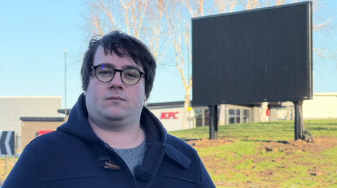 A man wearing black rimmed glasses and a dark blue jacket with a grey jumper stands in front of a roundabout where a 17ft high LED display has been installed. A branch of KFC can be seen in the distance.