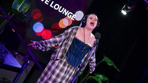 Lola Young sings in the Radio 1 Live Lounge studio. She wears a checked shirt and trousers, and a denim corset