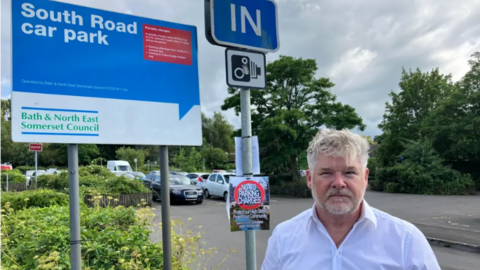 A photo of Independent Councillor Shaun Hughes, who is wearing a white shirt, with and open collar, standing in front of a car park in Midsomer Norton. There are various road signs next to him, one reading 'South Road car park'. 