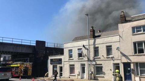 A large plume of grey smoke can be seen behind a building. Firefighters are reeling out hoses from a fire engine which is parked underneath a bridge.