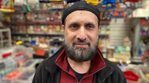 Shafiq Ahmed wearing a red jumper and black fleece and hat with his sweet shop blurred in the background