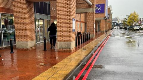 Double red lines painted outside shops on a retail park in Preston. You can see cars parked on the right hand side of the image, and shops including Currys on the left hand side. 