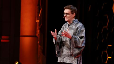 Llion Jones yn sefyll ar lwyfan gyda chefndir oren a phatrymau hecsagon du wedi’u gosod ar draws y wal. Mae’n gwisgo crys â phatrwm llwyd tywyll a golau, gyda llewys eang, ac mae ei ddwylo wedi’u codi wrth drafod gyda'r gynulleidfa.