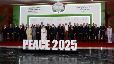 A row of people are stood on a red stage in front of white lettering that reads "PEACE 2025”.