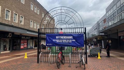 Wythenshawe's current shopping centre, made up of brown brick 1970s-style buildings. Shops including JD Sports and Select are under a dark canopy. A decorative black gate in the middle has a blue canvas sign on it that reads Wythenshawe: Big changes are coming that will make this place the place to be. Two bikes are locked up in front of the gate. 