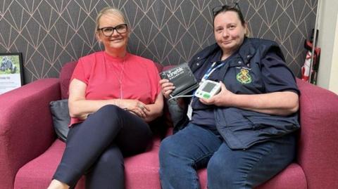 Two women are sitting on a pink sofa. The woman on the left has blonde hair,  black glasses and a pink top. Next to her, a woman has brown hair with black glasses on her head. She is wearing a navy first responder uniform, and is holding a blood pressure monitoring machine.