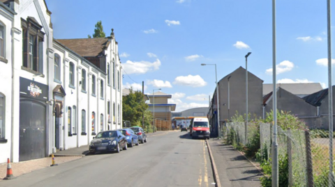 A number of parked cars parked along the side of a street with white buildings on one side and a metal fence on the other