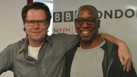 Robert Elms wearing a grey shirt over a white T-shirt, standing next to Ian Wright who is wearing a grey T-shirt an black jacket