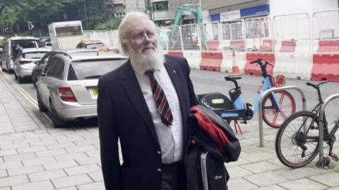 David Wright is pictured outside court. He is wearing a suit and tie and has a long white beard and glasses. He is carrying a bag and coat in his arms. Behind him are bicycles next to a rack and cars parked on the side of a road.