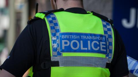 The back of a British Transport Police officer wearing uniform. He is wearing a black t-shirt and a yellow hi-visibility vest with blue and grey markings reading "British Transport Police" in capital letters.