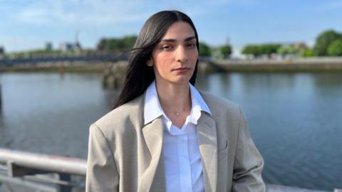 Ellie Wilson looking directly at the camera. She has long, dark hair. She is wearing a beige jacket over a white shirt and has a silver necklace around her neck. She is standing in front of a body of water and green foliage is blurred in the background. The sky abover her is light blue.