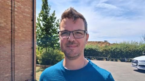Philip Wood stood in a car park, looking at the camera. He has short brown hair and facial hair and is wearing a blue T-shirt and black-rimmed, rectangular glasses. Behind him in the distance is a large pile of dirt on a construction site.