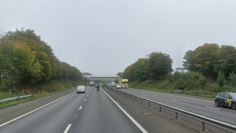 Vehicles drive on a motorway with trees either side. In the distance a building bridges across the road.